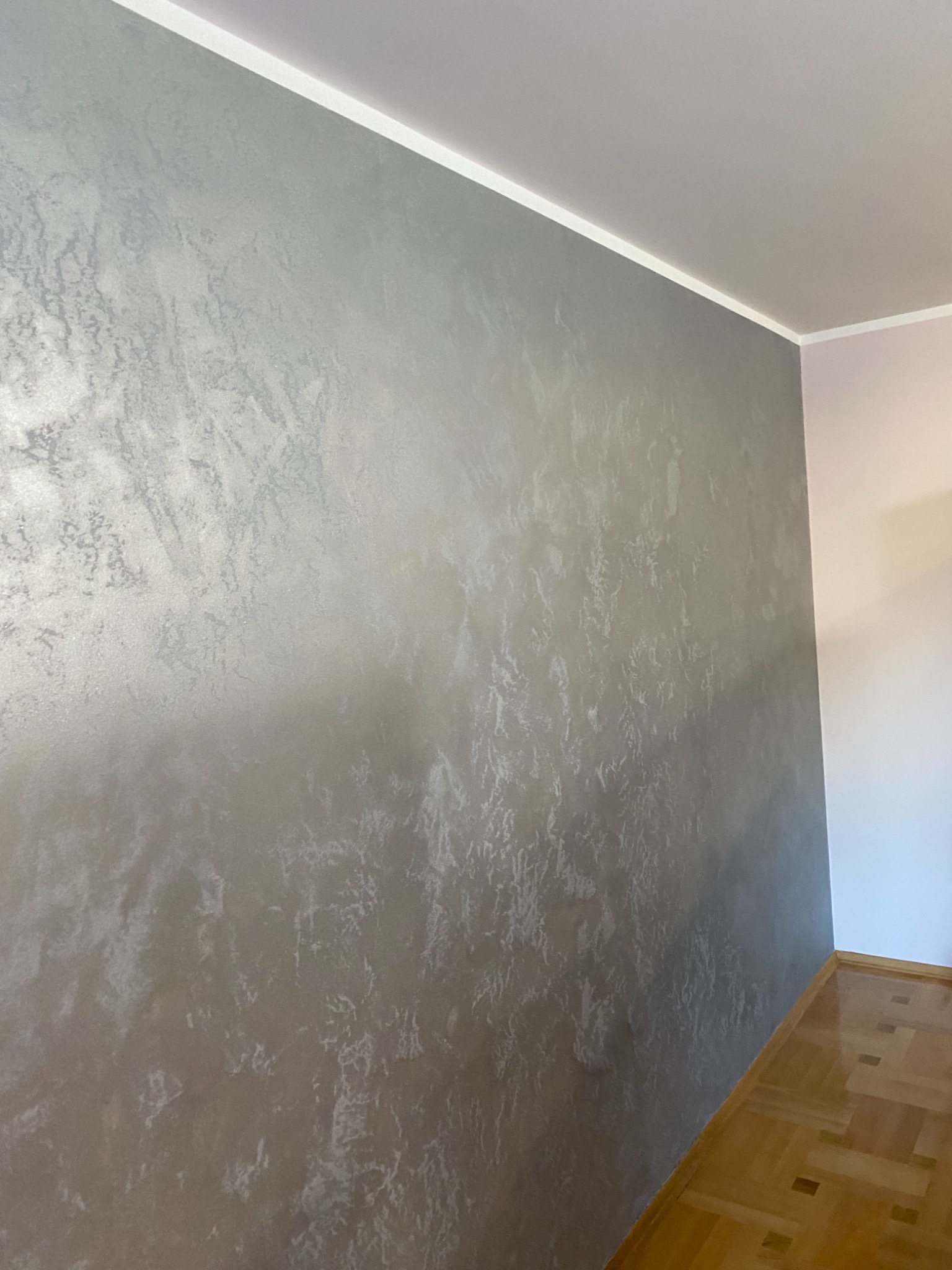 Ściana z dekoracyjną, srebrzystą fakturą, widoczna listwa przypodłogowa i fragment parkietu.