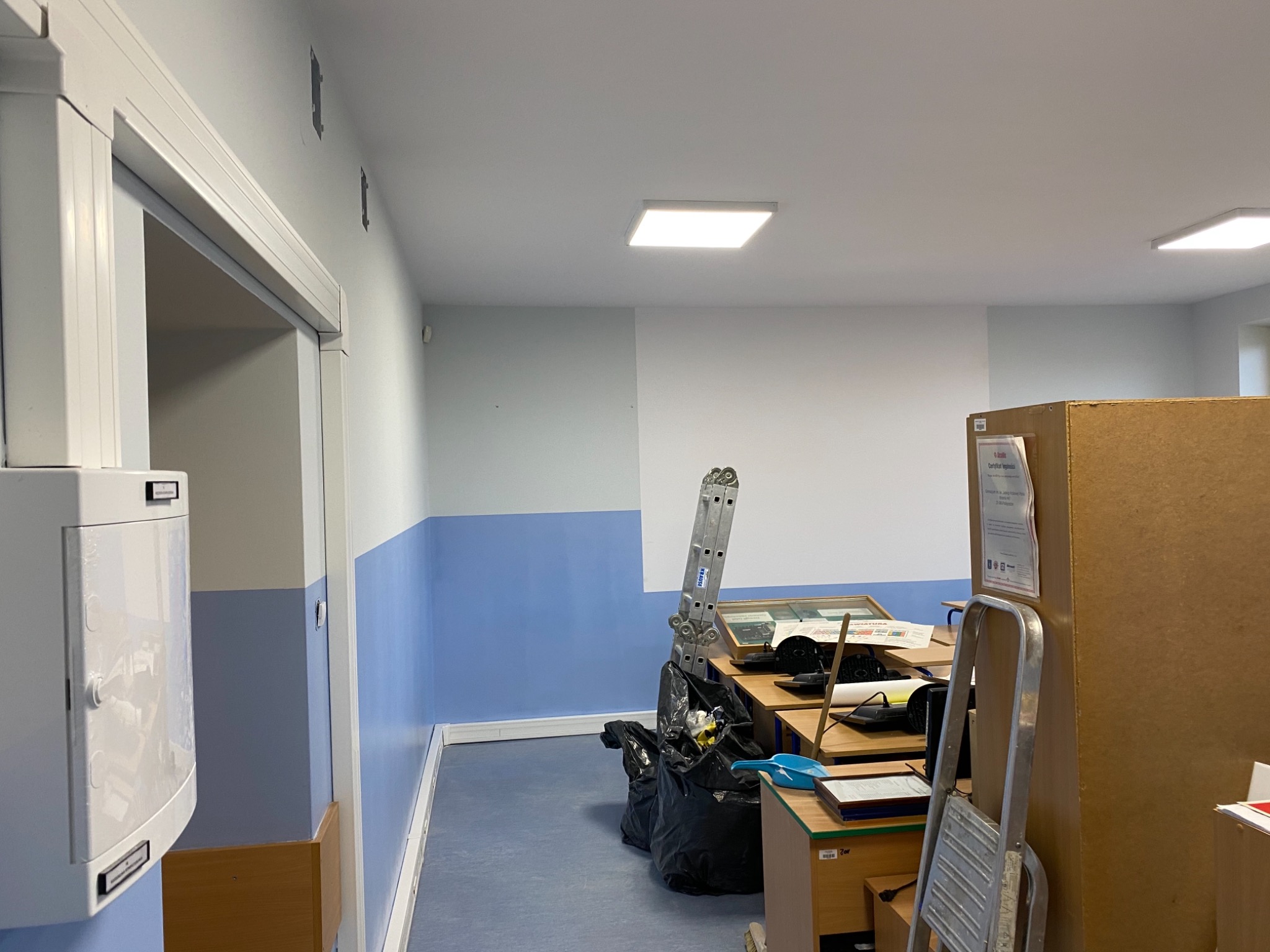 Wnętrze pomieszczenia z pomalowanymi ścianami na biało i niebiesko, widoczna drabina, biurka i worki na śmieci, sugerujące prace remontowe w toku.