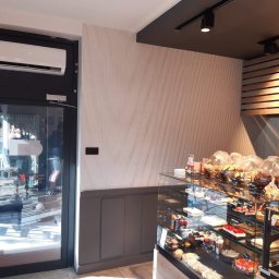 LeoKlima.pl - Biały klimatyzator zamontowany nad czarnymi drzwiami wejściowymi do lokalu, widok z wnętrza piekarni/cukierni z witryną pełną ciast i pieczywa.