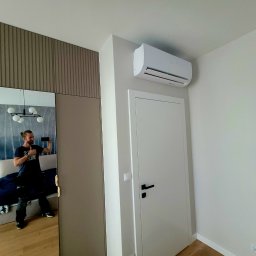 LeoKlima.pl - Biały klimatyzator ścienny zamontowany nad drzwiami w jasnym pokoju; w lustrze odbija się instalator robiący zdjęcie.