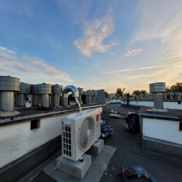 LeoKlima.pl - Biała jednostka zewnętrzna klimatyzacji LG Dual Inverter zamontowana na betonowych blokach na płaskim dachu z licznymi kominami wentylacyjnymi na tle błękitnego nieba o zachodzie słońca, obok...