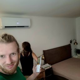 LeoKlima.pl - Uśmiechnięty mężczyzna trzyma pilot do klimatyzacji w sypialni z zamontowanym na ścianie klimatyzatorem, ustawionym na 25 stopni Celsjusza.