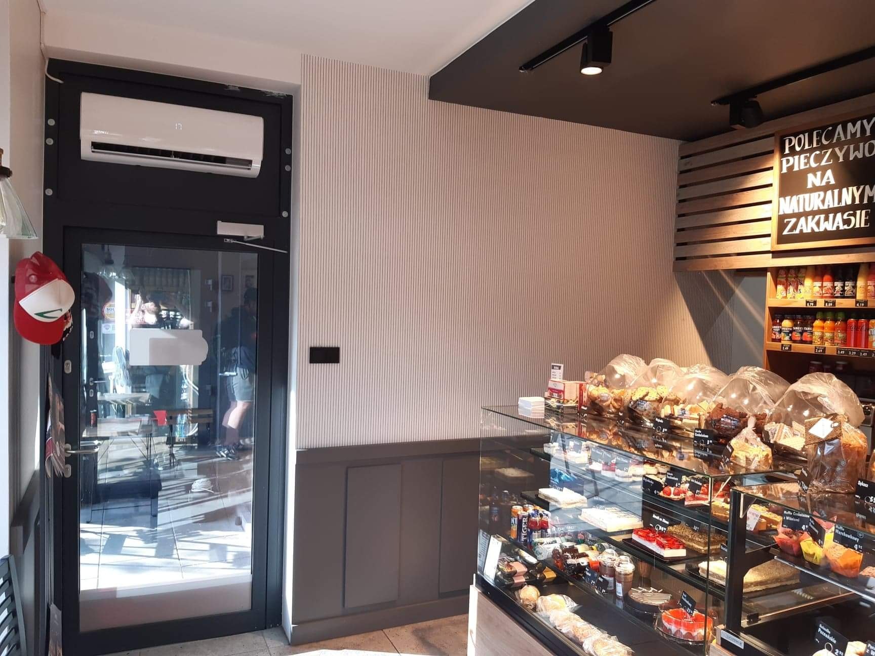 Biały klimatyzator zamontowany nad czarnymi drzwiami wejściowymi do lokalu, widok z wnętrza piekarni/cukierni z witryną pełną ciast i pieczywa.