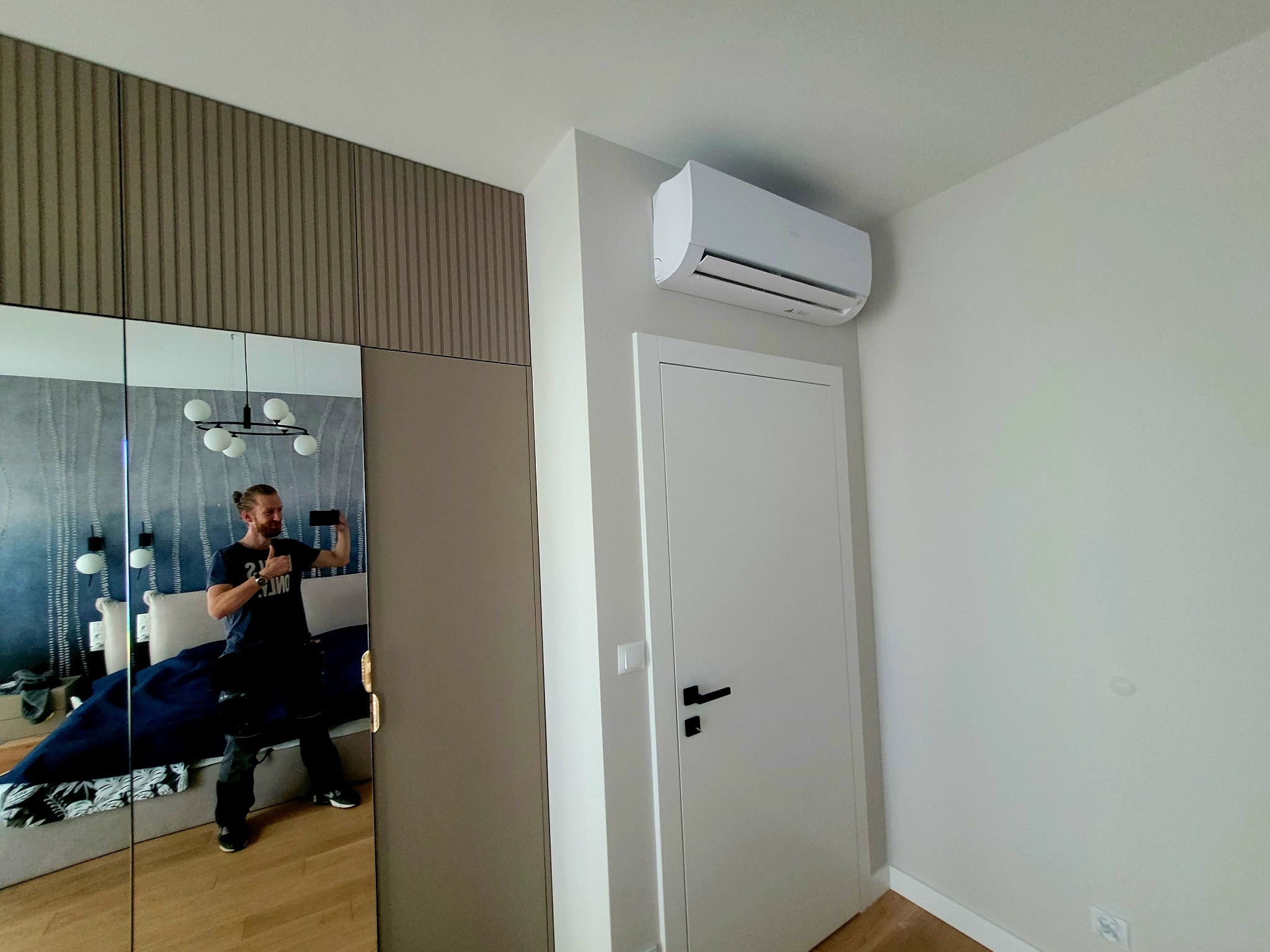 Biały klimatyzator ścienny zamontowany nad drzwiami w jasnym pokoju; w lustrze odbija się instalator robiący zdjęcie.
