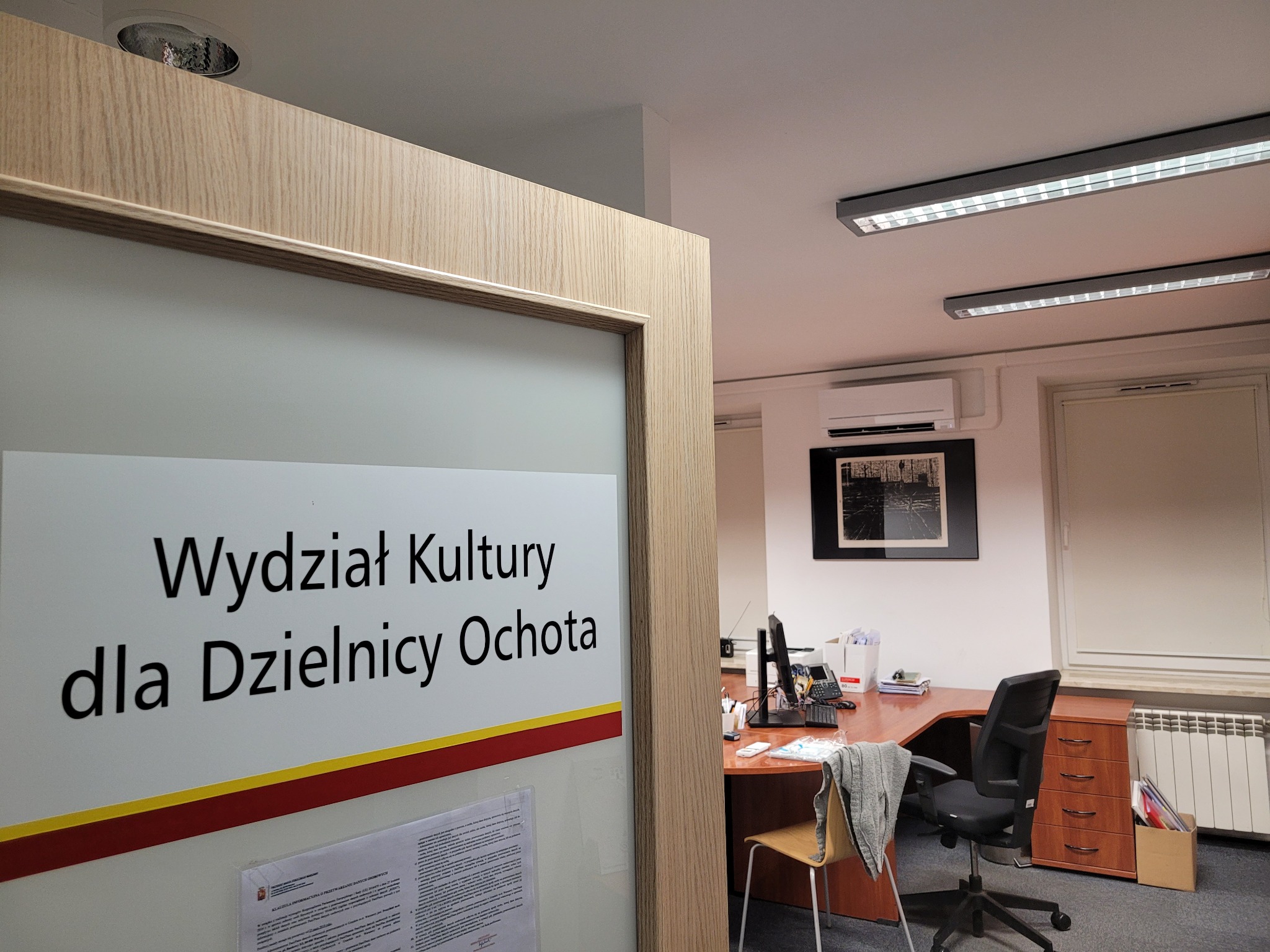 Wnętrze biura z zainstalowanym klimatyzatorem ściennym, widoczne zza przeszklonych drzwi z napisem 'Wydział Kultury dla Dzielnicy Ochota'.