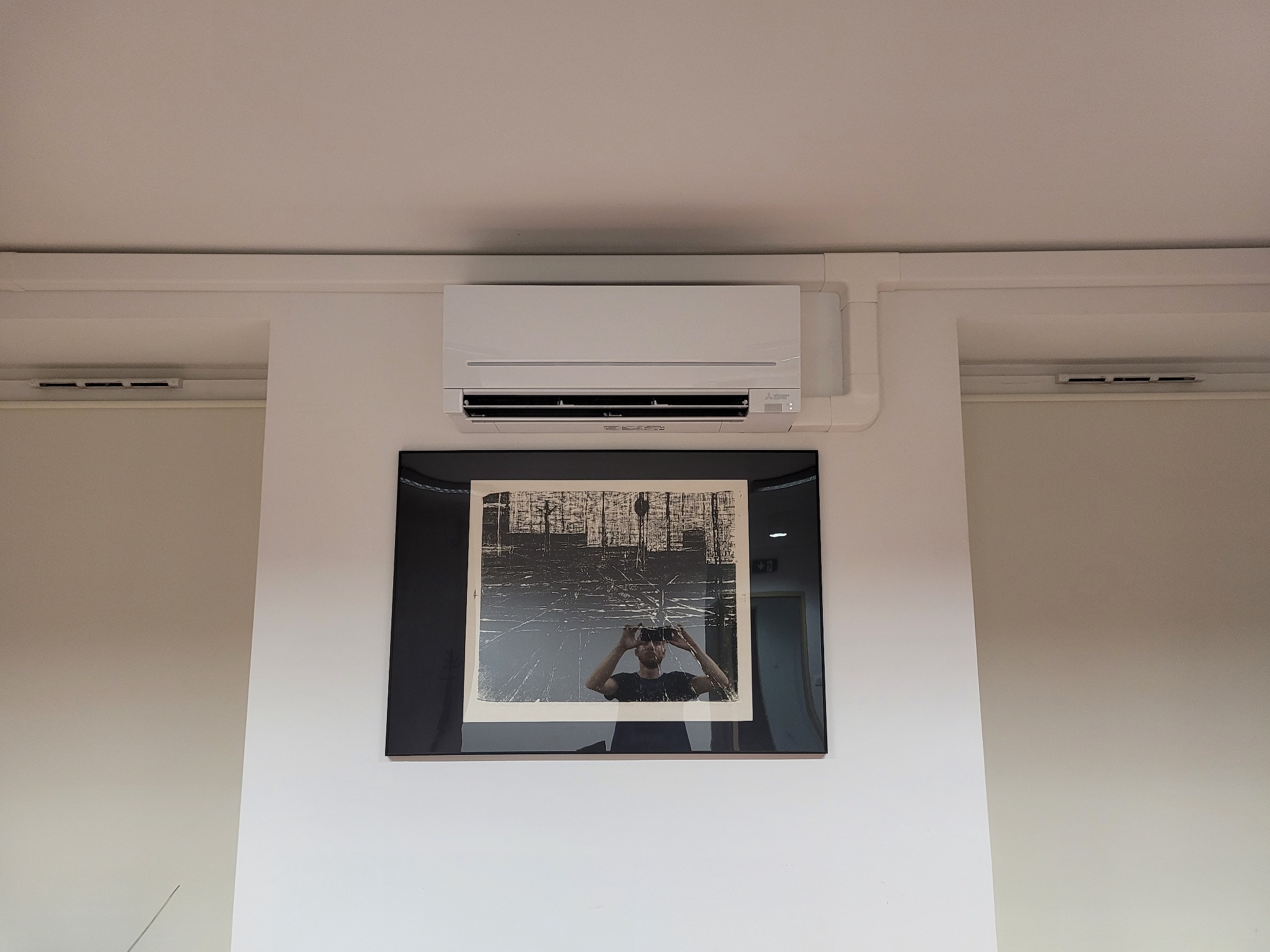 Biały klimatyzator ścienny zamontowany w pomieszczeniu z obrazem w czarnej ramie poniżej.