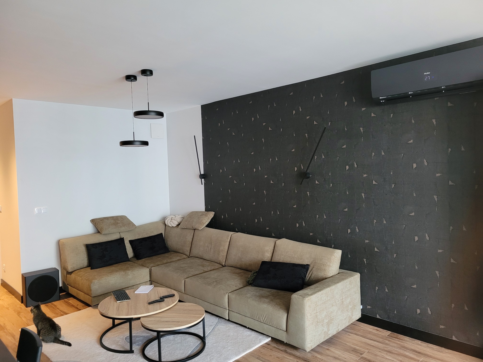 Nowoczesny salon z narożną sofą, okrągłymi stolikami kawowymi, wiszącymi lampami i zamontowaną na ścianie klimatyzacją Haier z wyświetlaczem temperatury, uzupełniony o subwoofer i ciekawskiego kota.