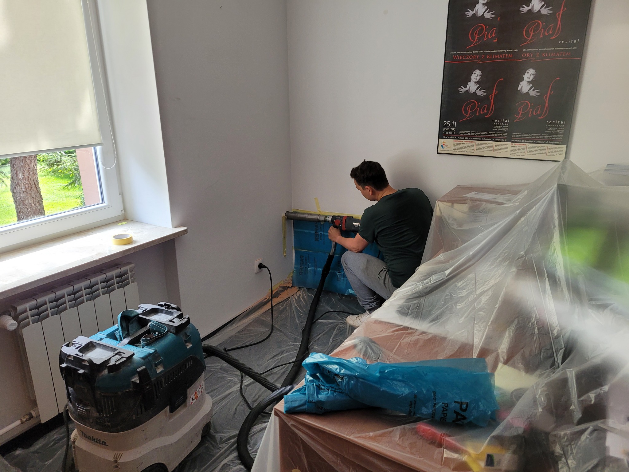 Instalator wierci otwór w ścianie pod rurę klimatyzacji w pomieszczeniu zabezpieczonym folią, w tle plakat teatralny z napisem 'Wieczory z klimatem'.