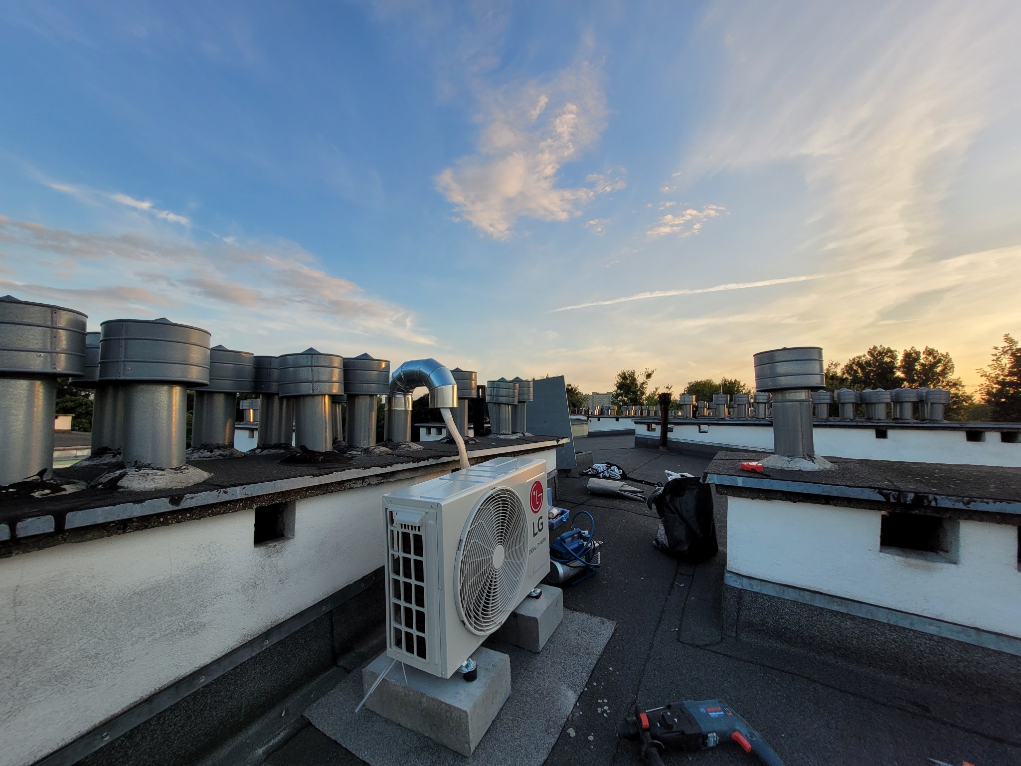 Biała jednostka zewnętrzna klimatyzacji LG Dual Inverter zamontowana na betonowych blokach na płaskim dachu z licznymi kominami wentylacyjnymi na tle błękitnego nieba o zachodzie słońca, obok...