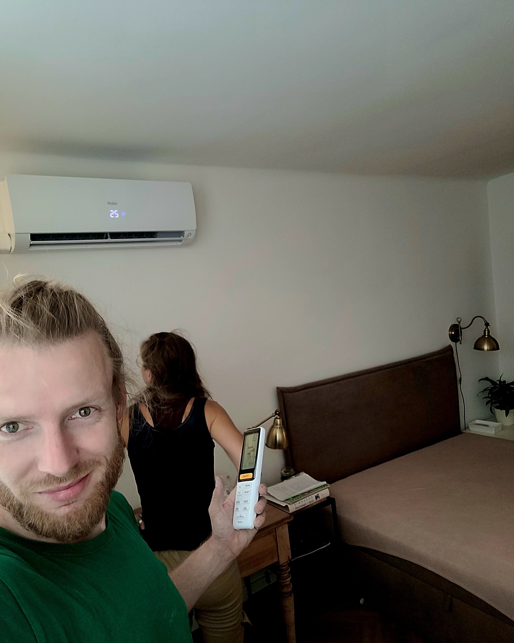 Uśmiechnięty mężczyzna trzyma pilot do klimatyzacji w sypialni z zamontowanym na ścianie klimatyzatorem, ustawionym na 25 stopni Celsjusza.