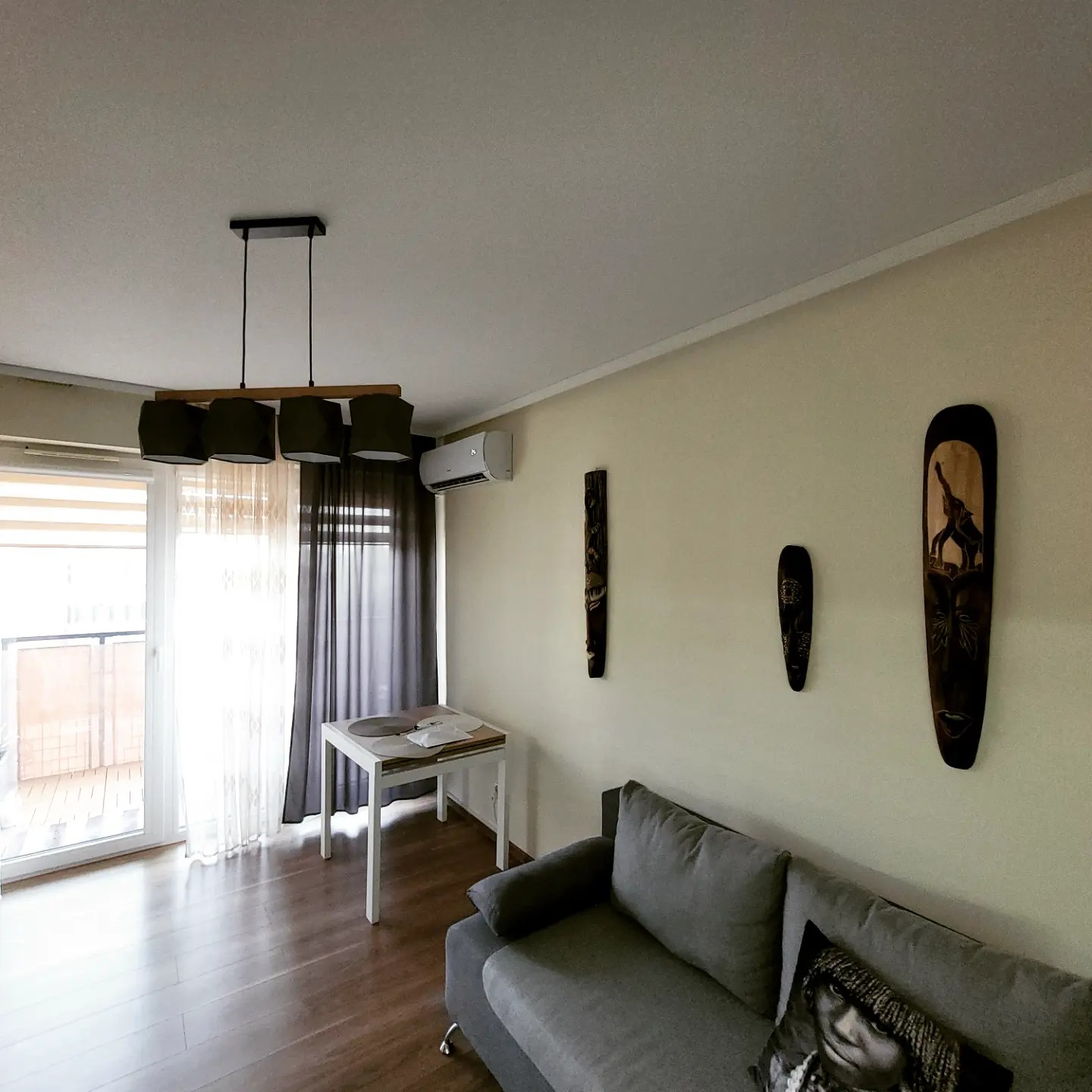 Jasne wnętrze salonu z zamontowaną klimatyzacją na ścianie, widoczne maski afrykańskie na ścianie, szara kanapa, stół i lampa wisząca.