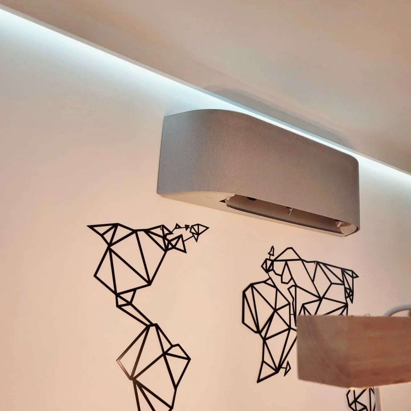 Szary klimatyzator ścienny z dekoracją w postaci geometrycznej mapy świata na białej ścianie, oświetlony listwą LED podsufitową.