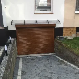 Podjazd z szarej kostki brukowej prowadzący do brązowej bramy garażowej rolowanej, zadaszonej przezroczystą osłoną. Oświetlenie podjazdu stanowią niskie, białe lampy. Widoczny fragment elewacji...