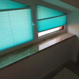 Turkusowe, plisowane osłony okienne częściowo zasłonięte na oknie pod skosem, z marmurowym parapetem w jasnym wnętrzu.