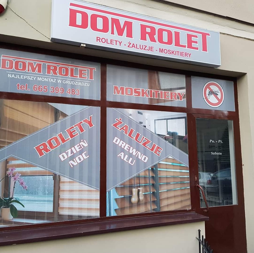 Fasada sklepu Dom-Rolet w Grudziądzu z ekspozycją produktów: okienne przesłony dzień noc, aluminiowe i drewniane konstrukcje, oraz siatki na owady, widoczne przez szybę z naklejonymi napisami i logo.
