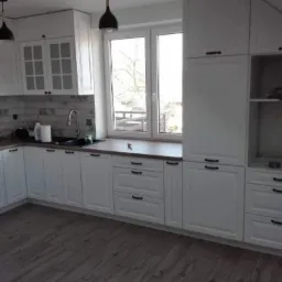 Jasna kuchnia z białymi szafkami i drewnianym blatem, czarne uchwyty i lampy, okno z widokiem na drzewa, podłoga z paneli imitujących drewno.