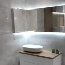 Nowoczesna łazienka z umywalką nablatową, drewnianym blatem i białymi szufladami, oświetlona listwą LED pod lustrem, na tle szarych płytek imitujących beton.