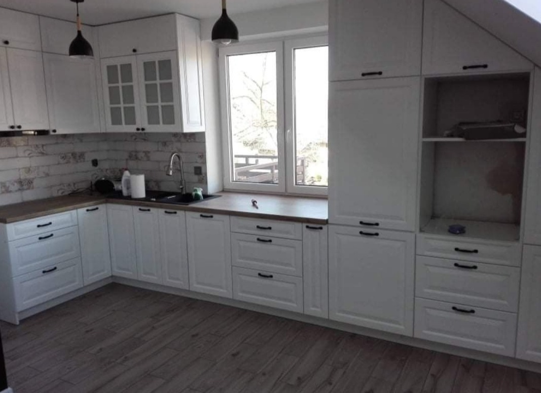 Jasna kuchnia z białymi szafkami i drewnianym blatem, czarne uchwyty i lampy, okno z widokiem na drzewa, podłoga z paneli imitujących drewno.