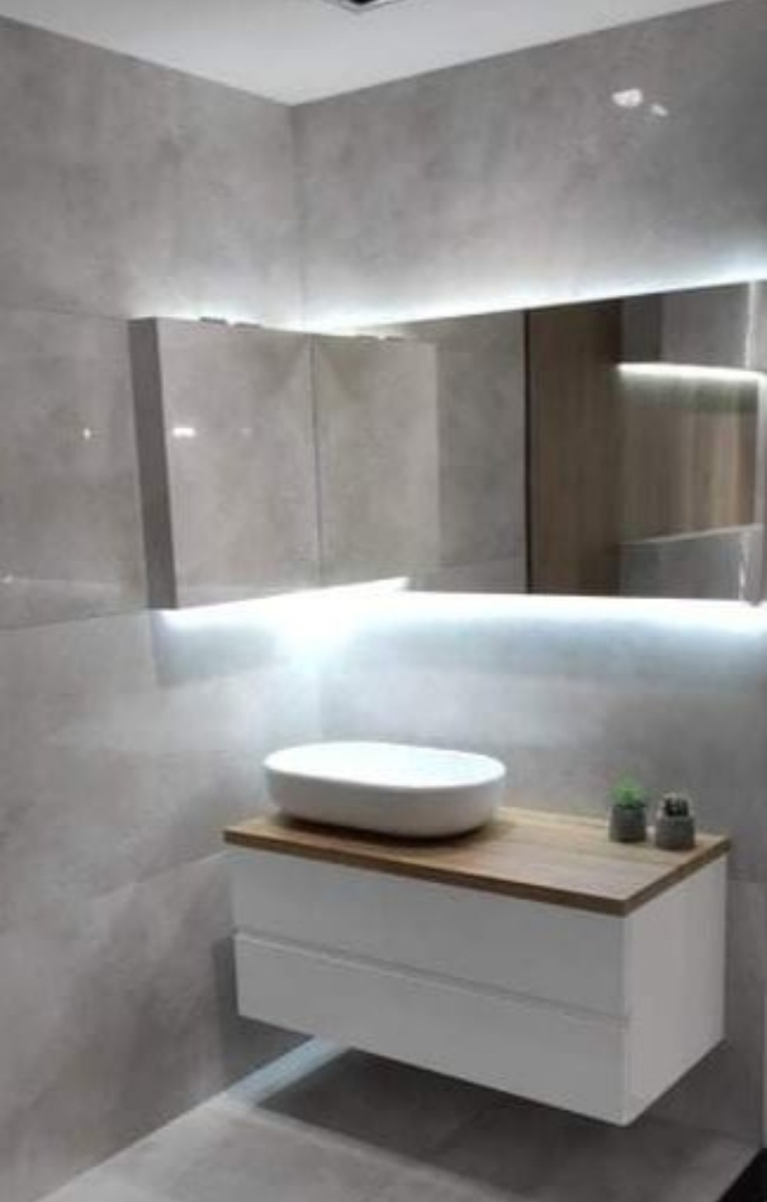 Nowoczesna łazienka z umywalką nablatową, drewnianym blatem i białymi szufladami, oświetlona listwą LED pod lustrem, na tle szarych płytek imitujących beton.