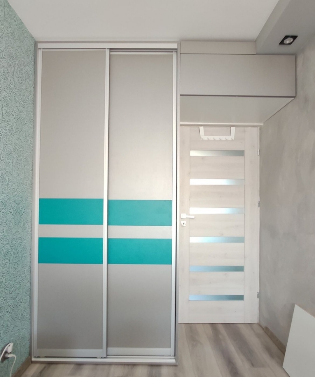 Dwudrzwiowa szafa przesuwna z poziomymi pasami w kolorze turkusowym, dopasowana do zabudowy wnęki, zintegrowana z szafką nad drzwiami i oświetleniem punktowym.