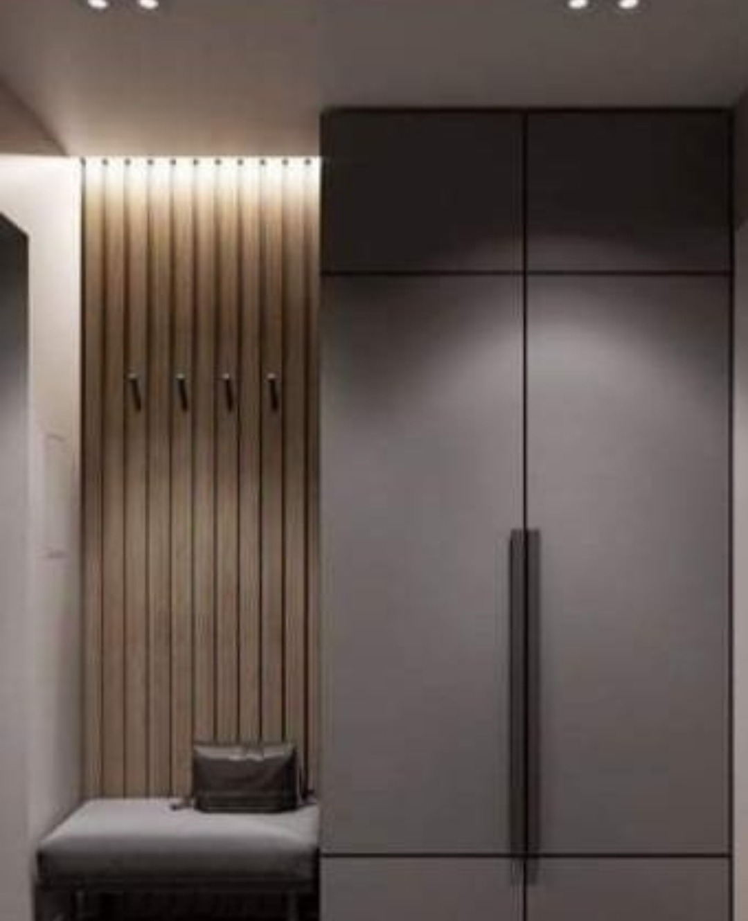 Nowoczesna szafa wnękowa w odcieniach szarości, z drewnianym panelem ściennym z wieszakami i siedziskiem z poduszką, oświetlona punktowo.