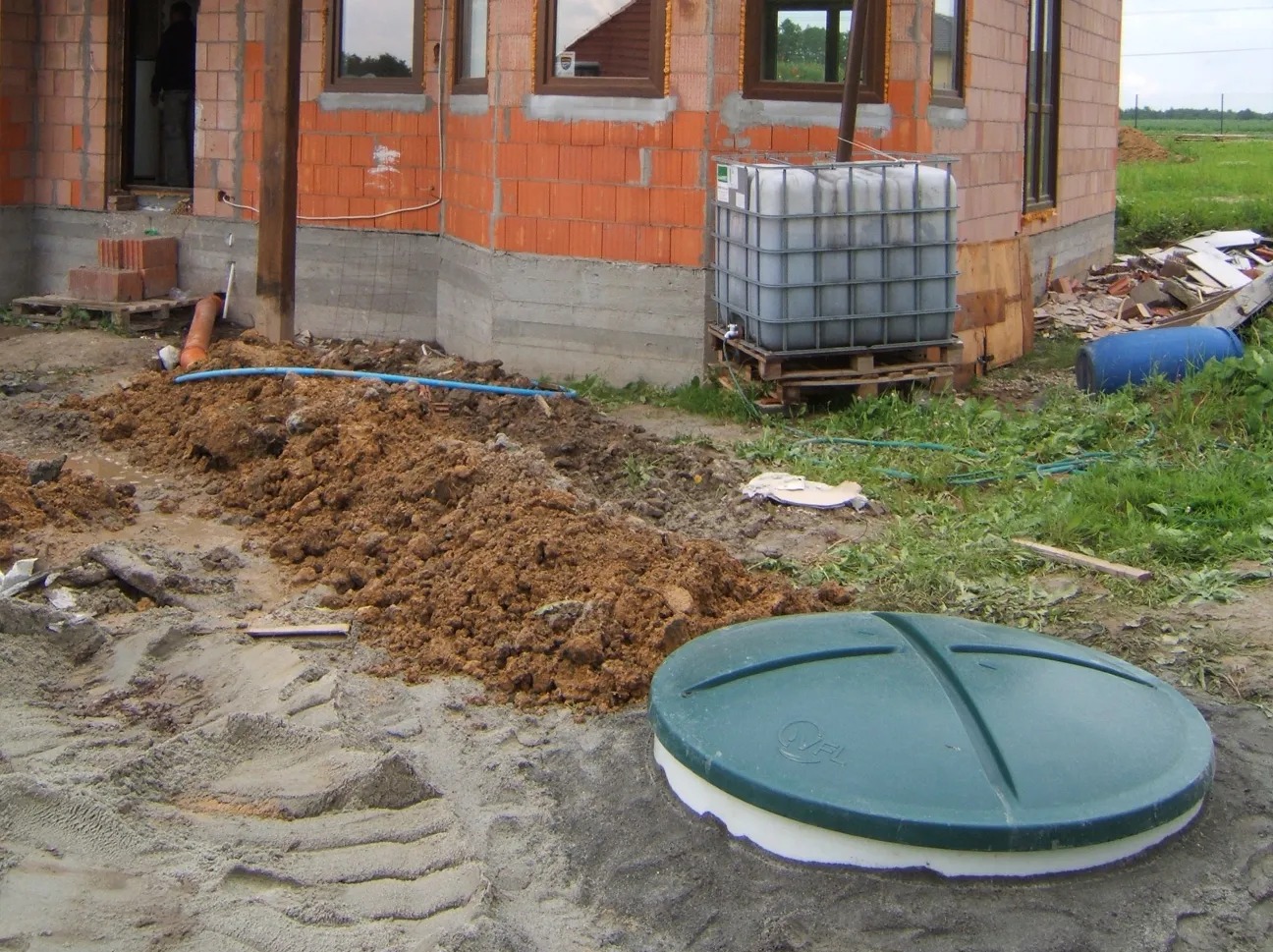 Montaż zielonej pokrywy szamba betonowego przy budynku z cegły. Widoczne rury, zbiornik na palecie i wykopana ziemia. Prace w toku.