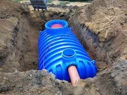 Niebieski zbiornik z tworzywa w wykopie, widoczny fragment koparki. Instalacja przydomowej oczyszczalni ścieków na etapie montażu. Ujęcie z góry, ziemia wokół.