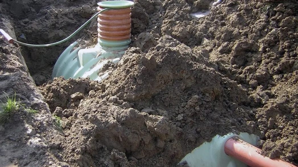 Montaż przydomowej oczyszczalni: zbiornik z zieloną pokrywą częściowo zakopany w ziemi, widoczne rury i wykop, ziemia usypana wokół zbiornika.