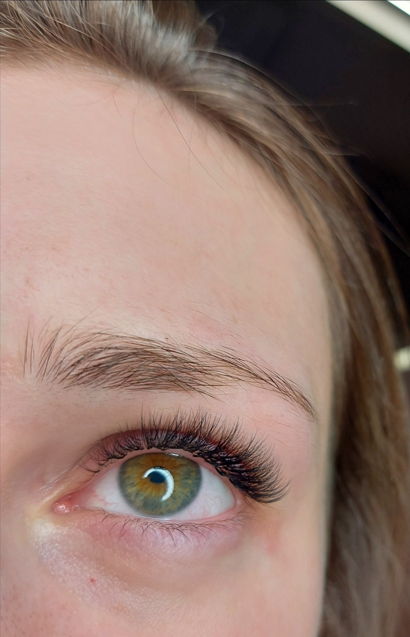 Zbliżenie na zielone oko z wyraźnie przedłużonymi, gęstymi rzęsami w stylu 'kocie oko', widoczna delikatna struktura brwi i jasna karnacja.