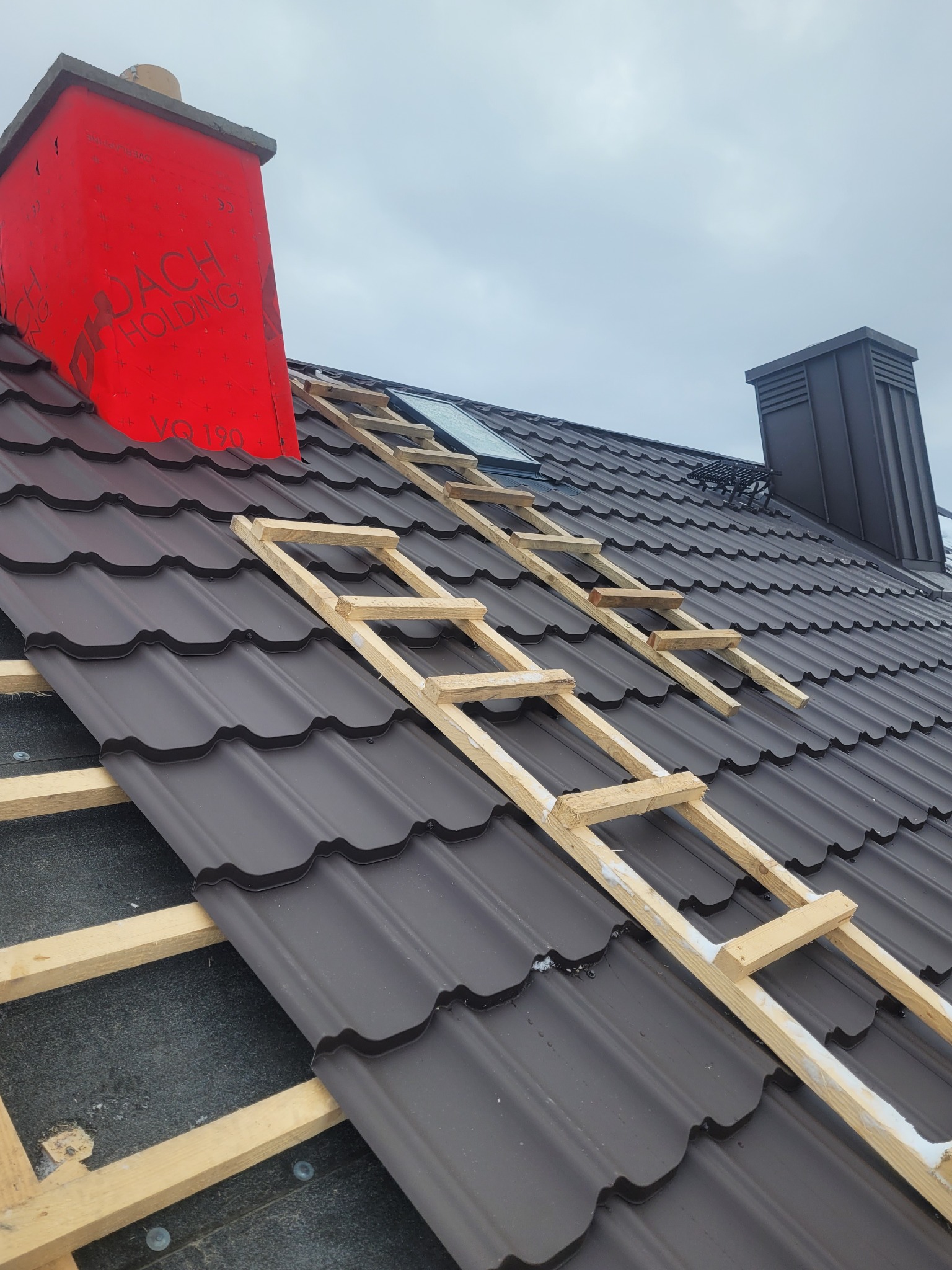 Montaż brązowej dachówki na dachu z czerwonym kominem w tle. Widoczne drewniane rusztowanie i fragment okna dachowego. Pochmurne niebo.