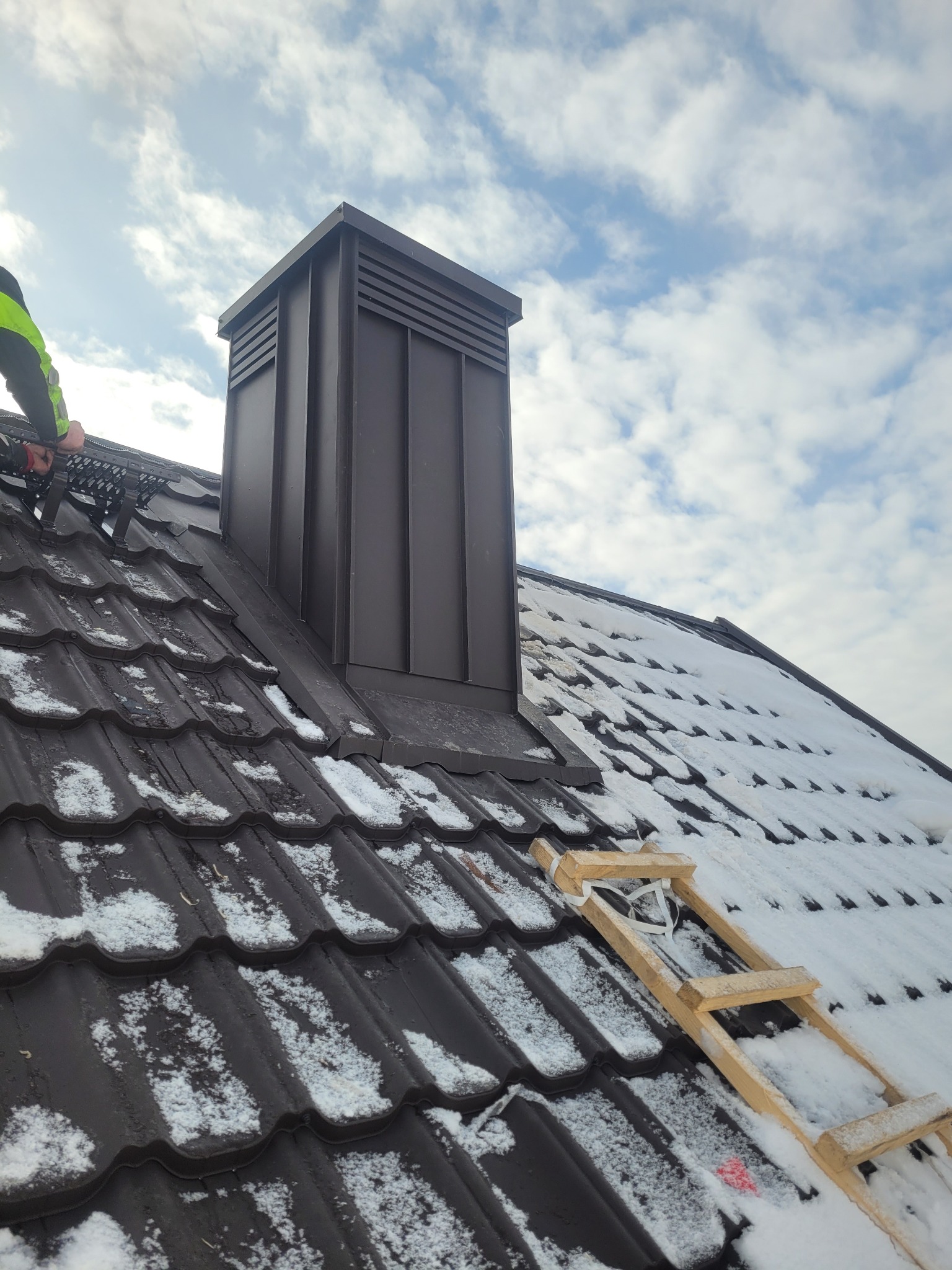 Dach z brązową dachówką i kominem w Szczytnie, częściowo pokryty śniegiem. Widoczny fragment drabiny dekarskiej i pracownik w odblaskowej kamizelce.