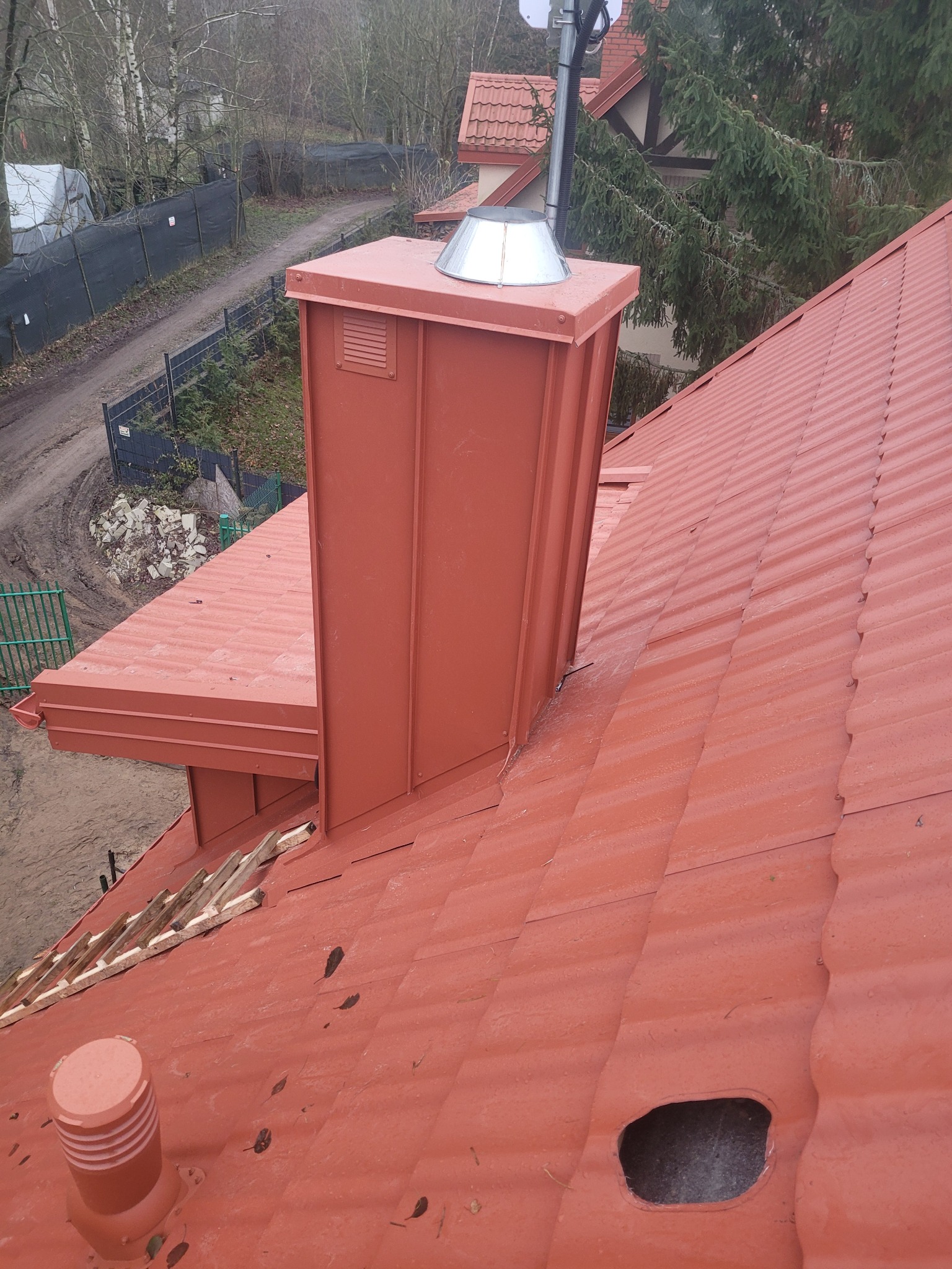 Czerwony dach z blachy trapezowej z kominem i elementami wentylacyjnymi. Widok z góry na ukończony dach w otoczeniu drzew i zabudowań.