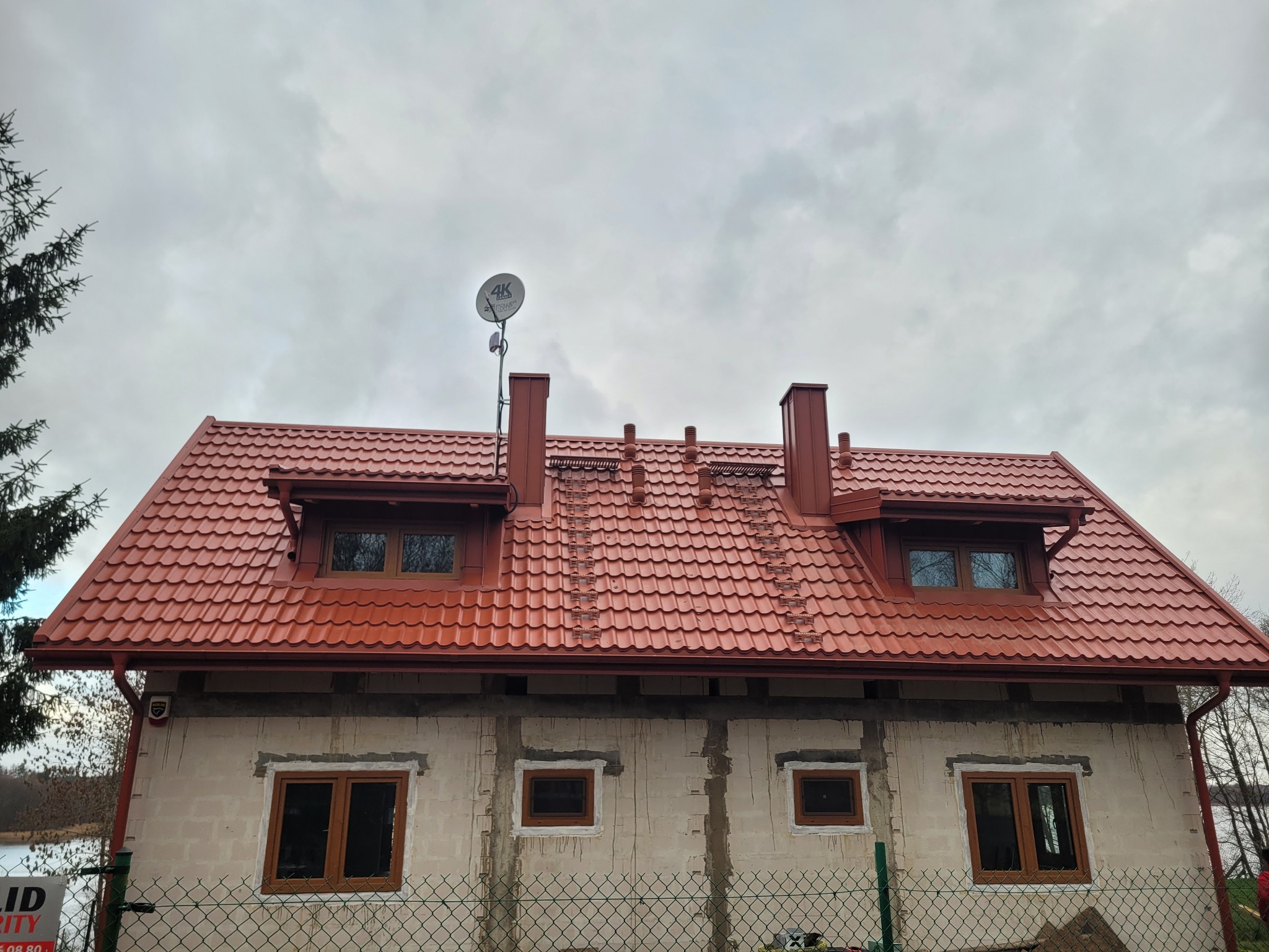Dom z nowym, czerwonym dachem z dachówki, widoczne kominy i antena satelitarna. Budynek w trakcie budowy, ściany nieotynkowane. Pochmurne niebo w tle.