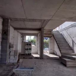 Wnętrze budynku w stanie surowym otwartym z widocznymi ścianami z betonu komórkowego, betonowym stropem, schodami oraz stertą bloczków H+H i taczką budowlaną.