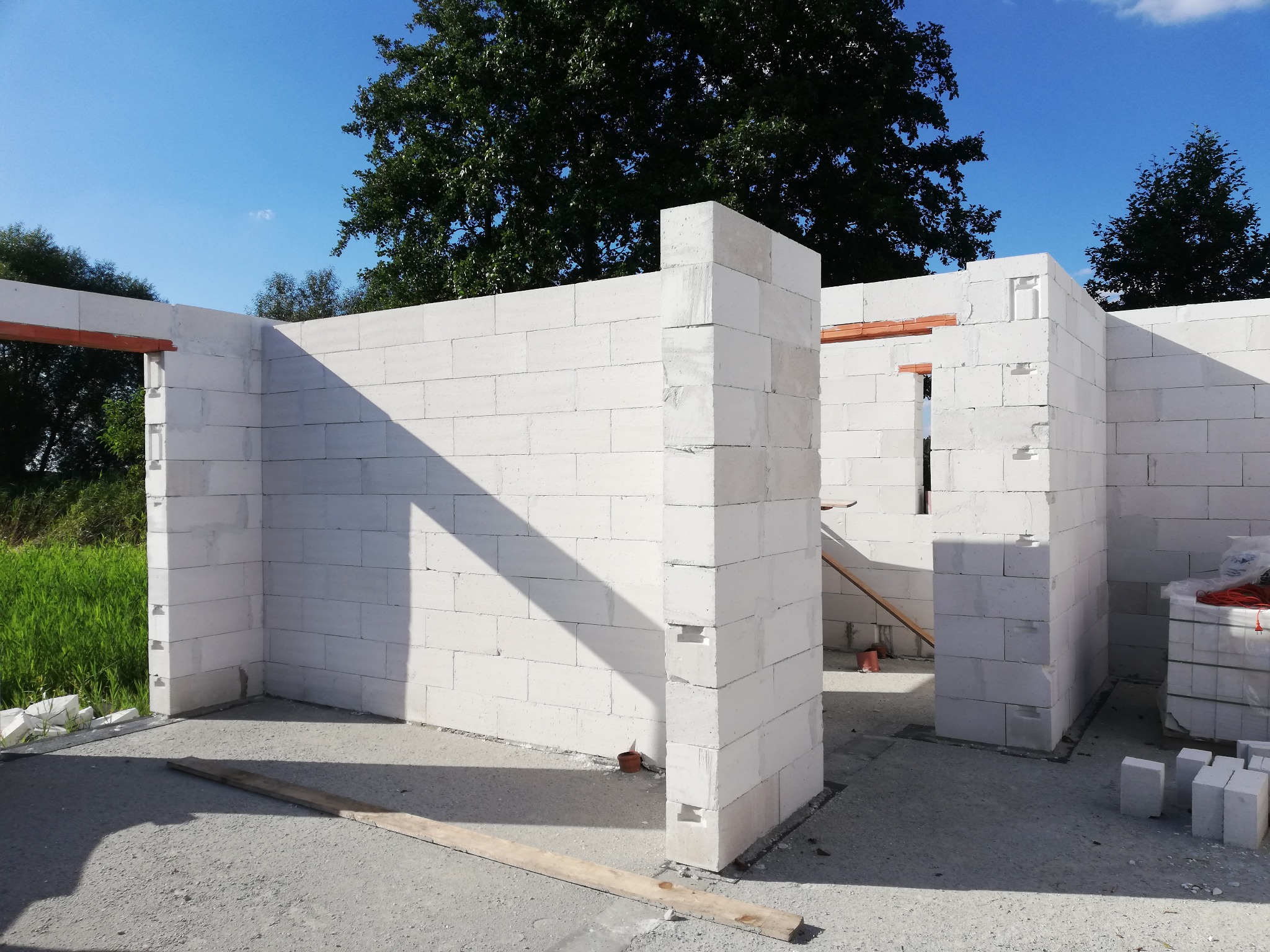 Wznoszenie ścian nośnych z bloczków Ytong na fundamencie, widoczne otwory okienne i drzwiowe, w tle zieleń i błękitne niebo.