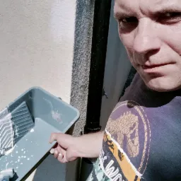 Mężczyzna w koszulce z motywem Jurassic World trzyma kuwetę malarską z resztkami białej farby, widoczna fragment elewacji budynku z rynną.