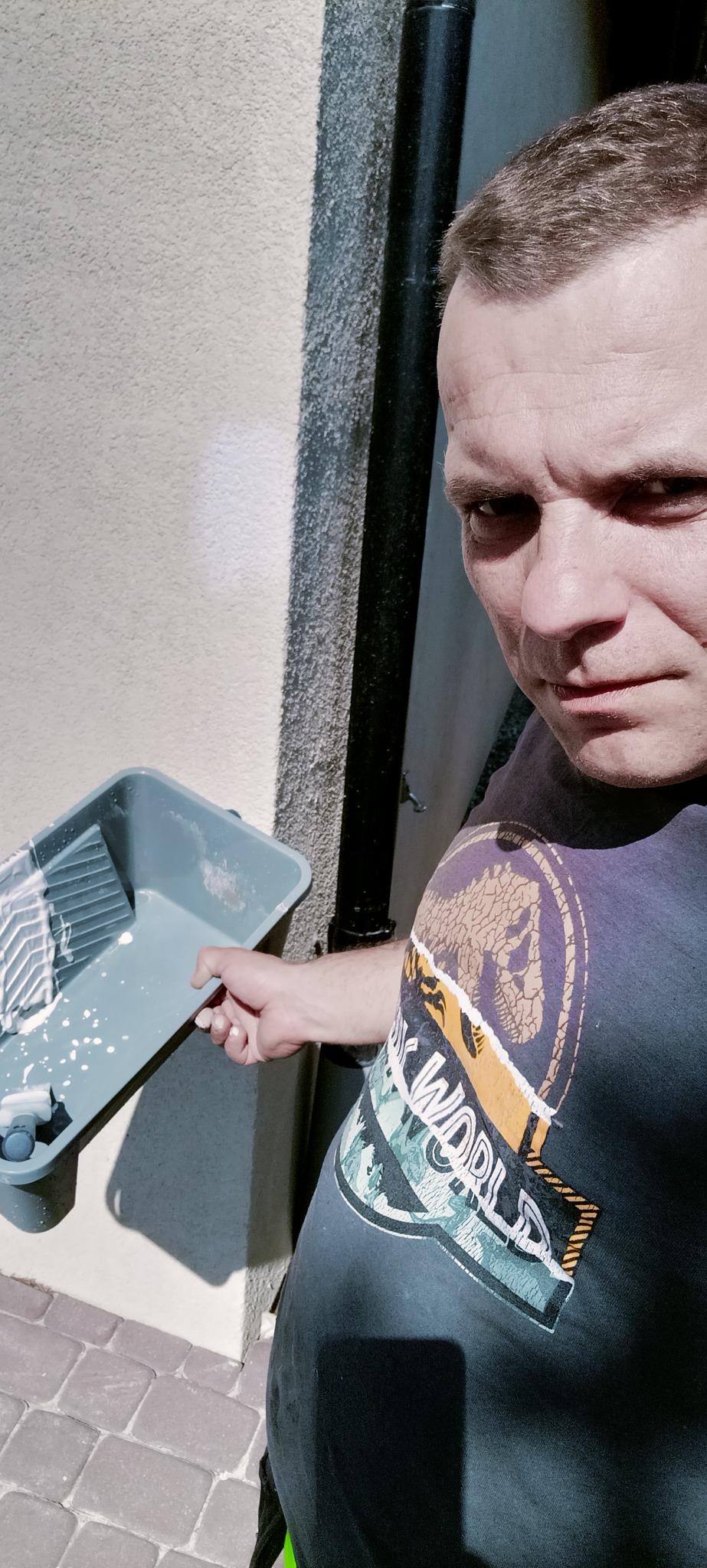 Mężczyzna w koszulce z motywem Jurassic World trzyma kuwetę malarską z resztkami białej farby, widoczna fragment elewacji budynku z rynną.