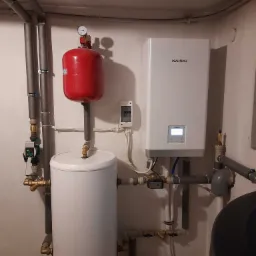 Zainstalowana biała pompa ciepła Kaisai, z czerwonym zbiornikiem wyrównawczym i białym zasobnikiem ciepłej wody w kotłowni z szarymi rurami instalacji grzewczej.