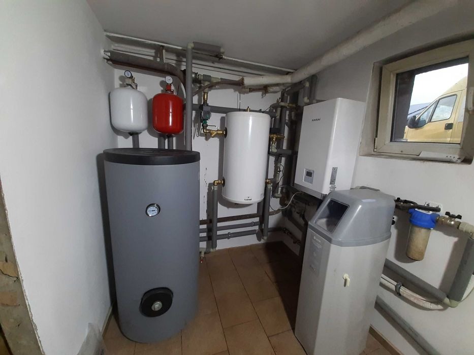 Instalacja grzewcza w piwnicy: szary zasobnik ciepłej wody z manometrem, biały zbiornik wyrównawczy, czerwony zbiornik ciśnieniowy, wiszący biały piec gazowy i filtr wody na tle białych ścian...