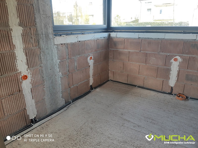 Surowy stan budynku z instalacją elektryczną: widoczne przewody w peszlach biegnące wzdłuż ścian z cegły ceramicznej, puszki elektryczne zalane gipsem, okno w tle.