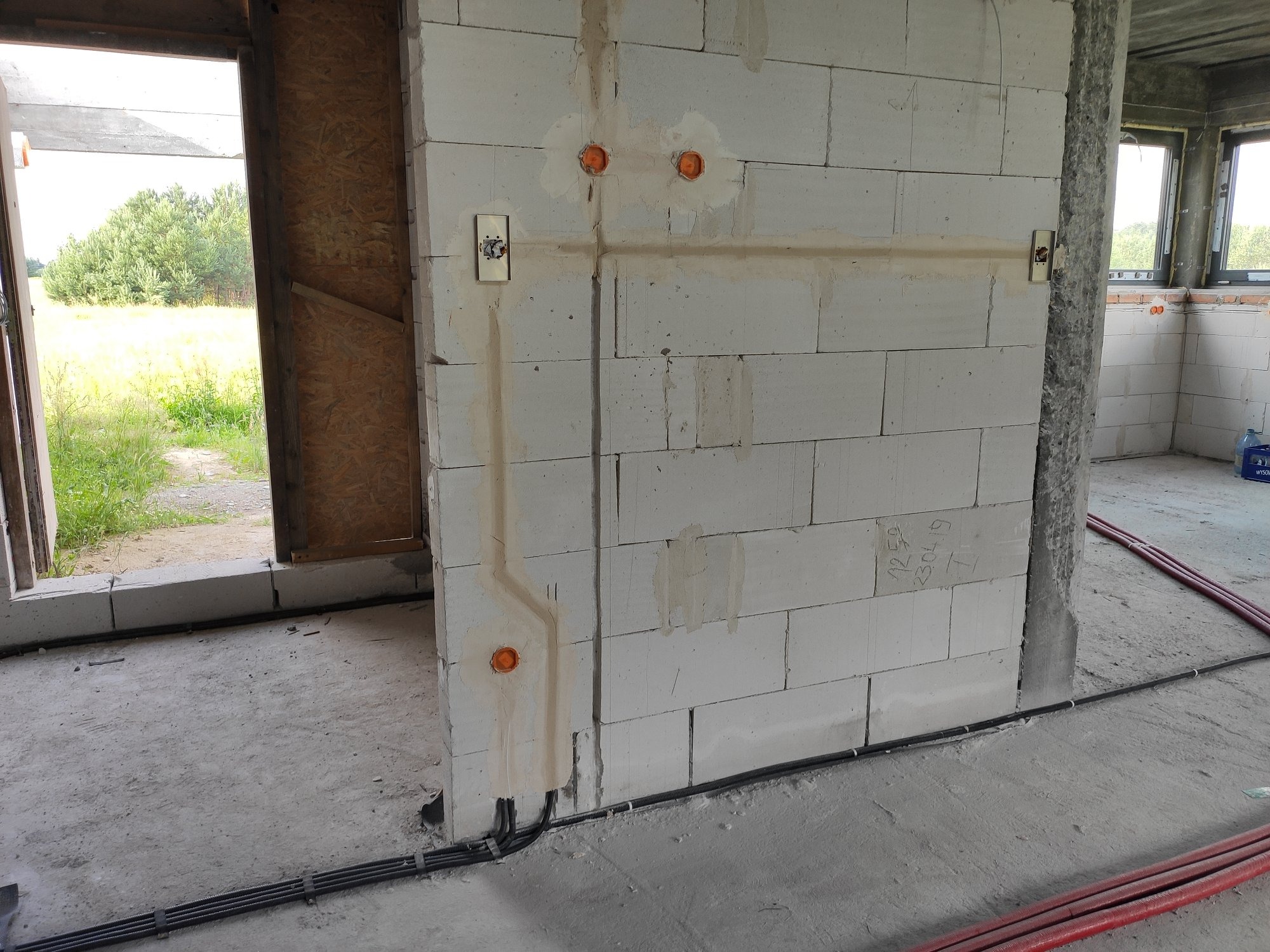 Wewnętrzna ściana z betonu komórkowego w trakcie budowy, widoczne bruzdy pod przewody elektryczne, puszki instalacyjne i częściowo zamontowany przełącznik światła. Na podłodze leżą rury...