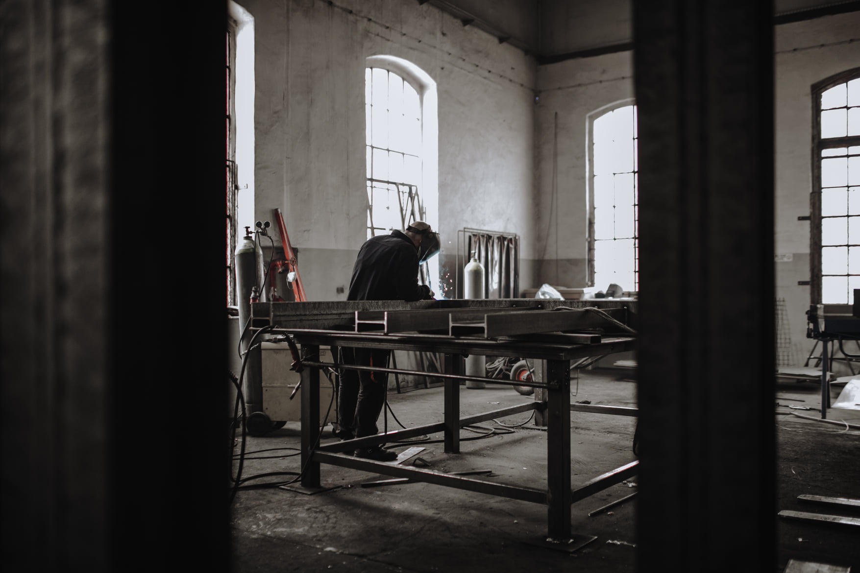 Spawanie metalowej konstrukcji w warsztacie, widoczne iskry i spawacz w masce ochronnej, butle z gazem w tle, surowe wnętrze z dużymi oknami.