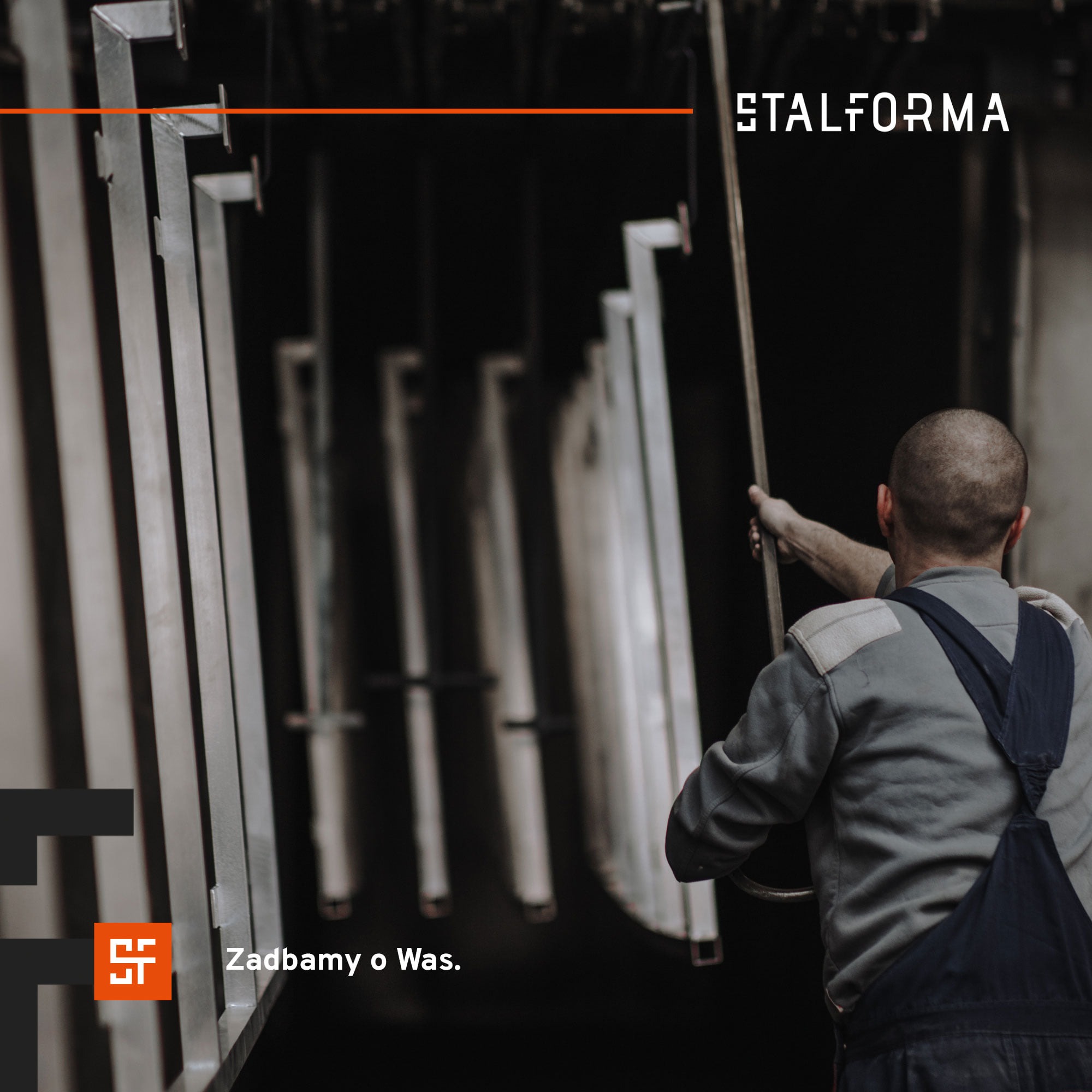 Pracownik w kombinezonie zawiesza metalowe elementy na wieszakach w hali produkcyjnej, logo firmy w lewym górnym rogu.