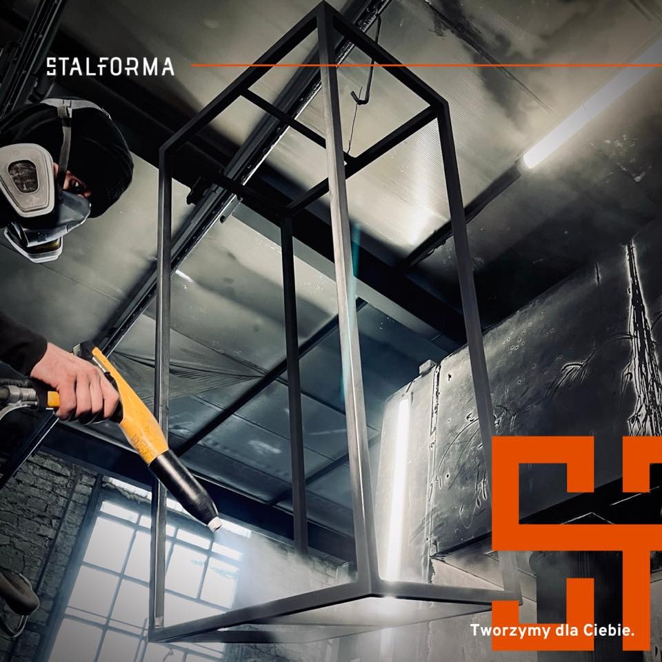 Malowanie proszkowe metalowej konstrukcji w kształcie prostopadłościanu przez pracownika w masce ochronnej. W tle ceglany mur i logo firmy Stalforma.