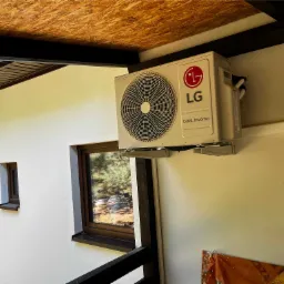 Zewnętrzna jednostka klimatyzatora LG Dual Inverter zamontowana na ścianie budynku z widocznymi oknami i drewnianym zadaszeniem.