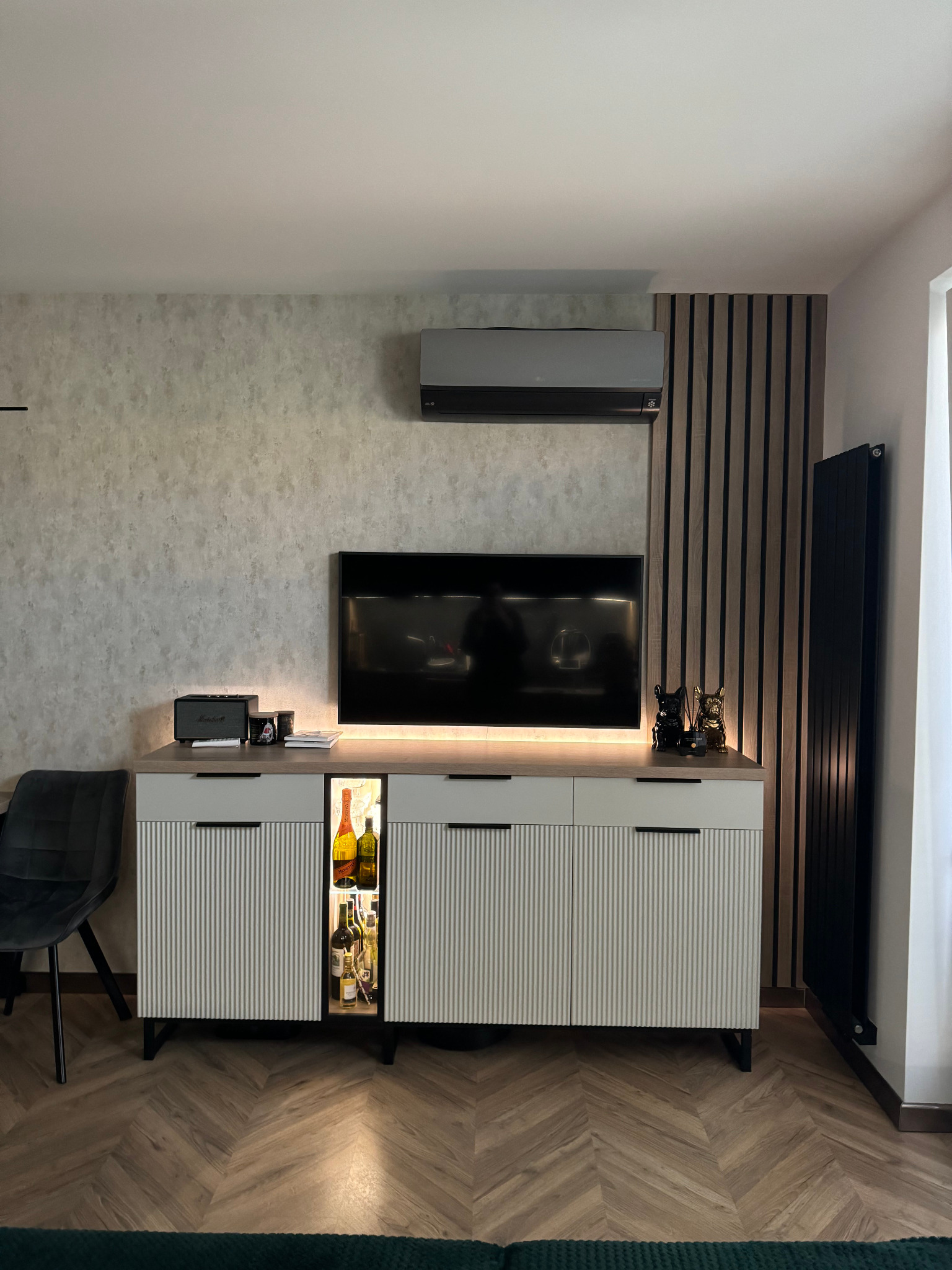 Nowoczesny salon z klimatyzatorem ściennym, szafką RTV z podświetlaną wnęką na napoje, dekoracyjną ścianką z lameli i grzejnikiem żeberkowym.