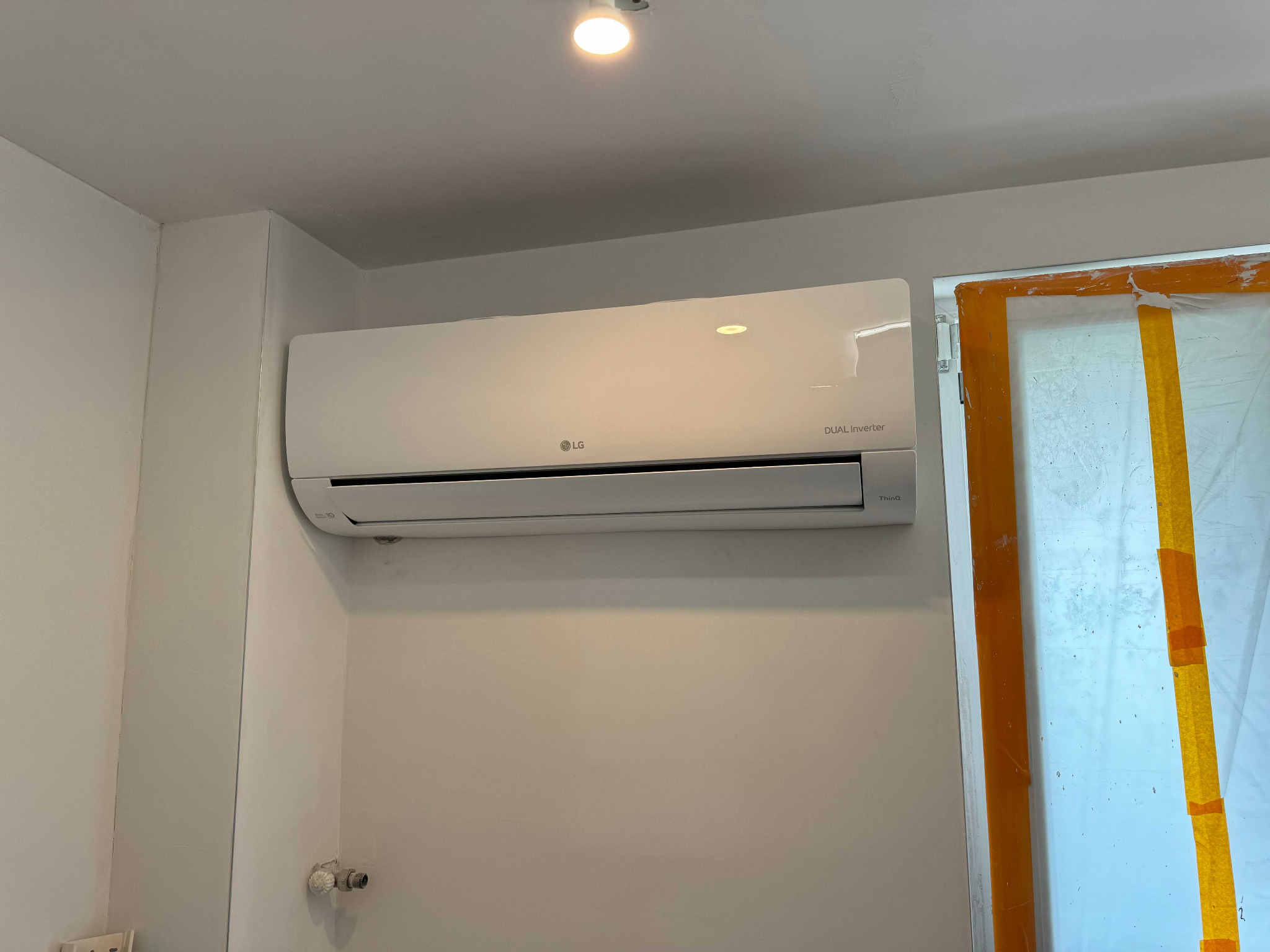 Biała jednostka wewnętrzna klimatyzatora LG Dual Inverter zamontowana na białej ścianie, widoczne zabezpieczone okno folią i taśmą malarską.