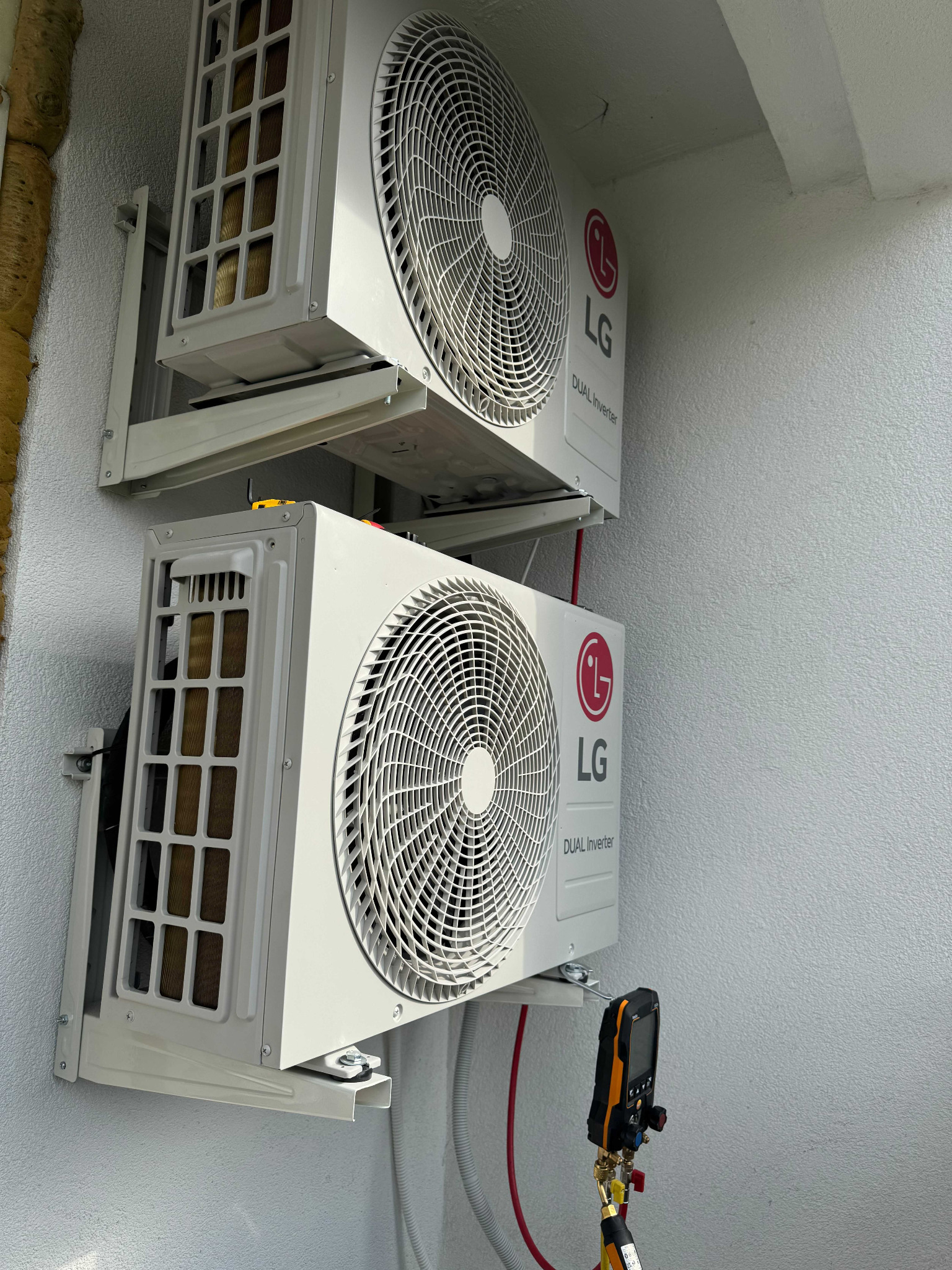 Dwa białe klimatyzatory LG Dual Inverter zamontowane jeden nad drugim na białej ścianie, widoczne narzędzie pomiarowe z czerwonym przewodem podłączone do dolnego klimatyzatora.