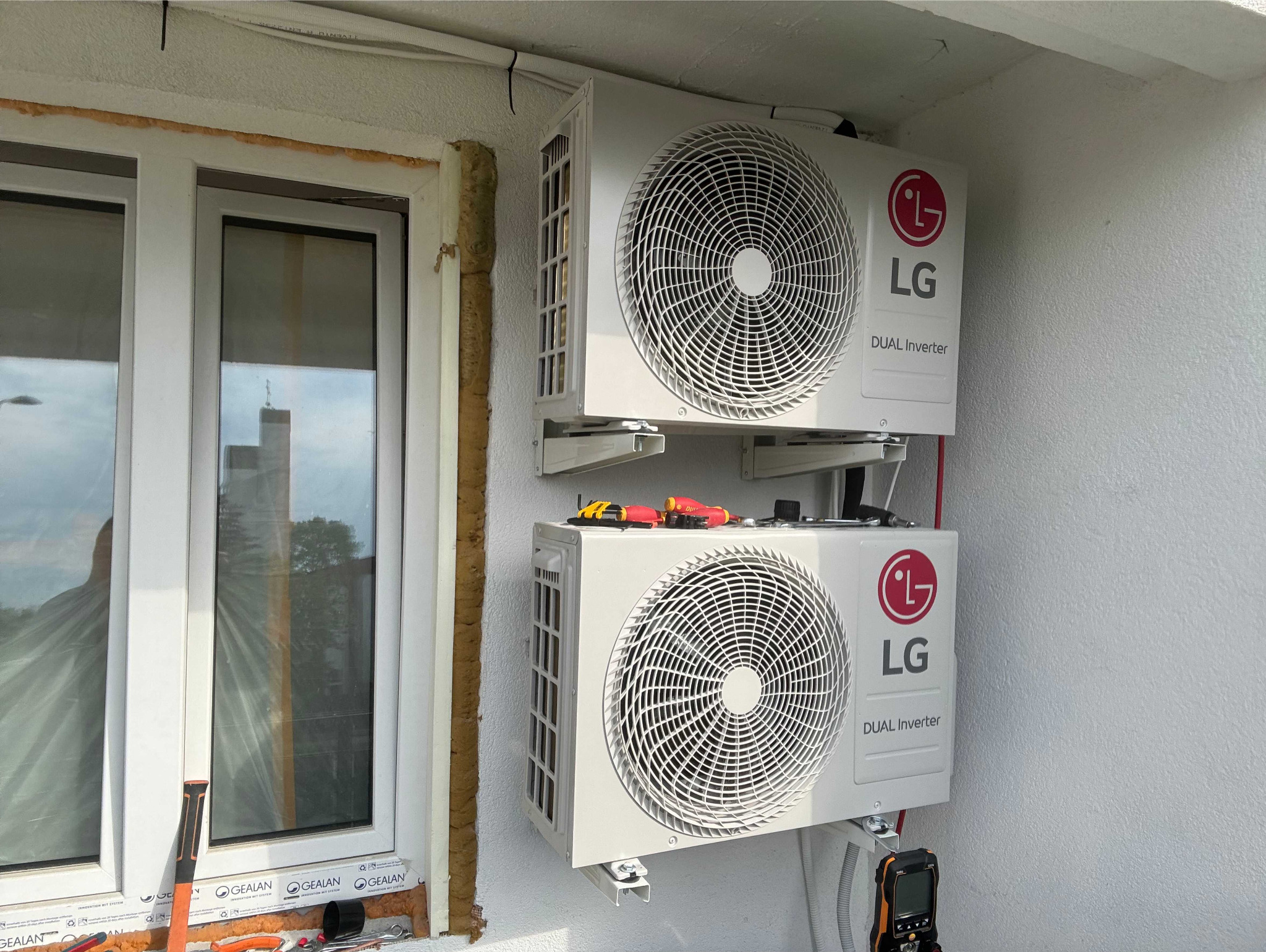 Dwa zamontowane na ścianie, białe klimatyzatory LG Dual Inverter, z narzędziami na górnym urządzeniu, obok okna z widocznym odbiciem budynku.