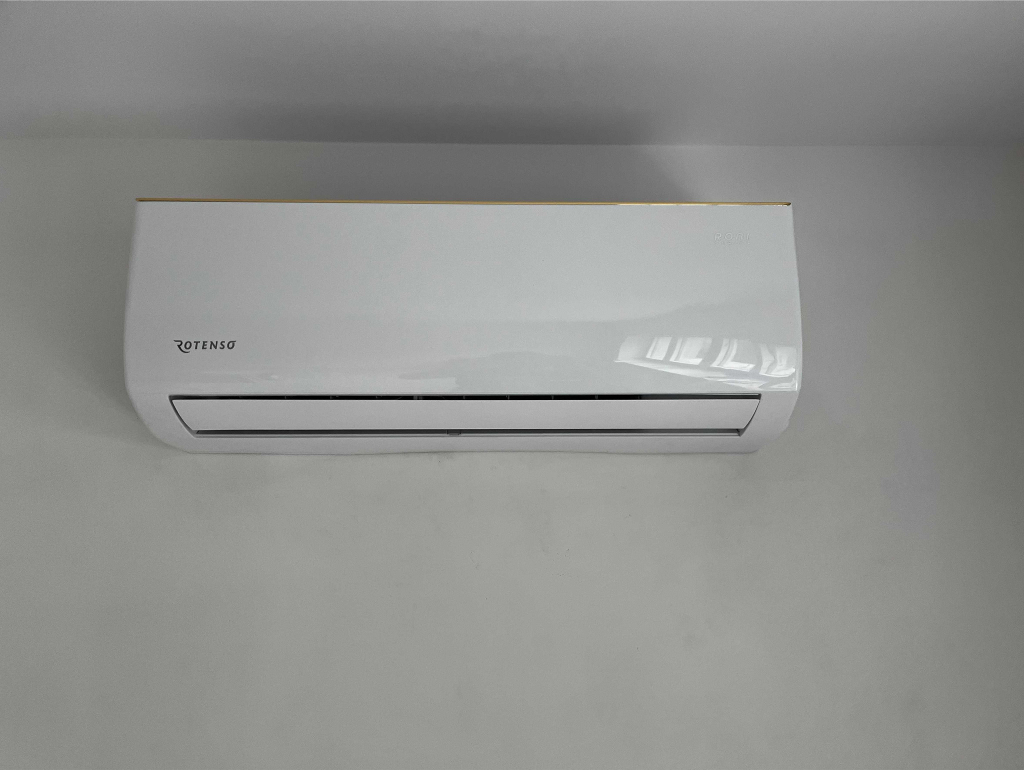 Biała jednostka klimatyzacji marki Rotenso zamontowana na szarej ścianie, z delikatnym złotym akcentem na górnej krawędzi.